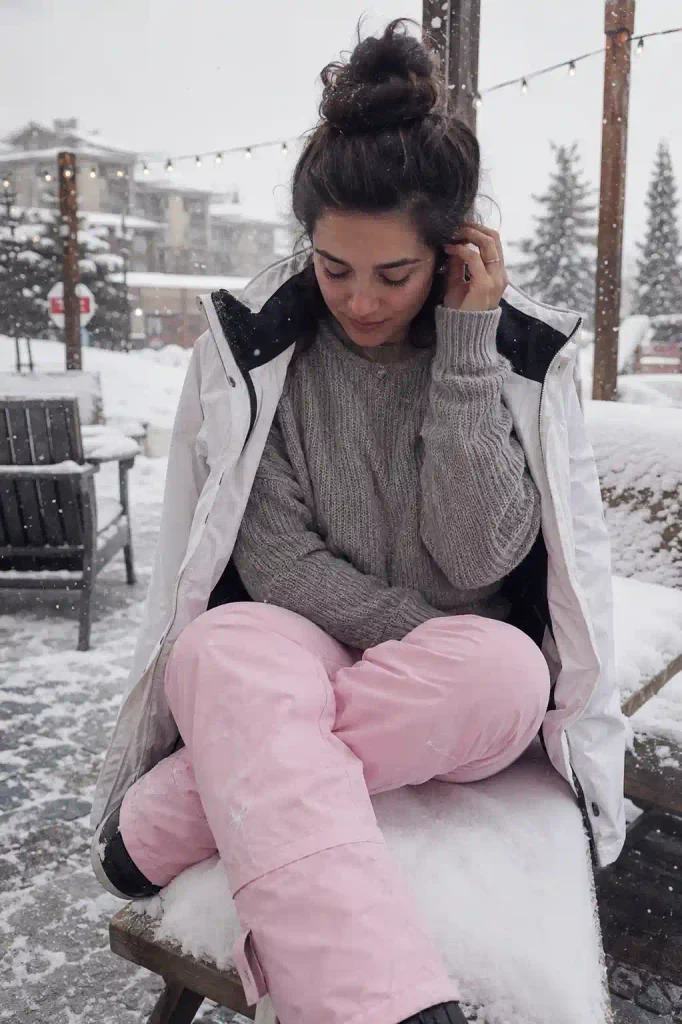 A woman sits on the tailgate of a car in a resort parking lot, one ski boot off and resting beside her, adjusting the cuff of a thin grey base layer visible where her pink ski pants are pulled up slightly at the ankle. The base layer fabric is thin and clingy against her skin, with natural wrinkling where it bunches near her ankle. The dusty rose ski pants pooled slightly at her calf show soft, heavy folds typical of technical shell fabric gathered rather than worn straight. Her posture is unglamorous and real, hunched slightly forward, focused on her own hands as she tucks the fabric into her sock, not looking at the camera at all. Her fingers are natural and slender, caught mid-motion. Her expression is neutral, a little furrowed in concentration, mouth slightly pressed, completely unposed. The shot is framed from the front but slightly to the side, close enough to see the layering detail clearly, with an imperfect, candid tilt as if caught in the middle of getting ready. Bright, cool midday light falls evenly across the parking lot snow, with a real shadow cast by the open car door beside her. Background shows a snowy parking area, other cars with roof-mounted ski racks, and a distant tree line. Natural grain, no studio lighting.