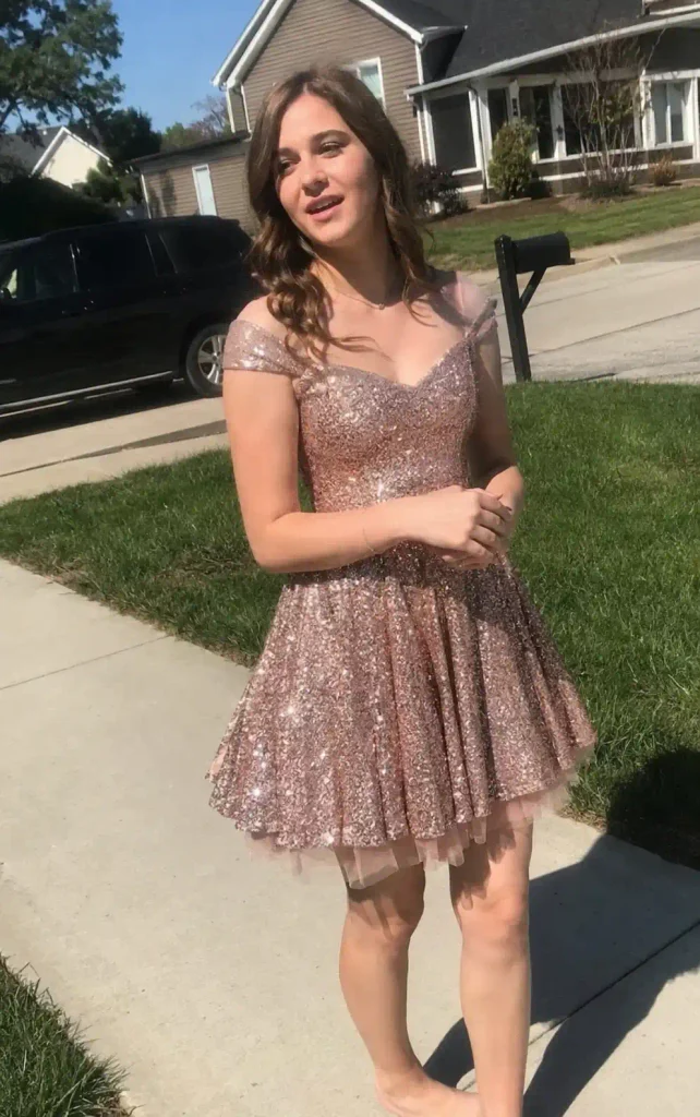 An adult American woman with a natural, believable figure stands near a wooden backyard fence in soft overcast daylight, wearing a rose gold satin slip dress with thin metallic shimmer threads woven through the fabric. The satin drapes loosely and catches diffuse light unevenly, with natural creasing near the hip where her hand rests. She is caught mid-laugh, head tilted slightly, with one hand adjusting a thin strap on her shoulder. Her skin shows natural texture and a faint blemish near her jaw, with no airbrushing or excessive retouching. Behind her, a softly blurred garden, lawn chair, and tall grass sway gently. The scene has the soft, cool light of an overcast afternoon, subtle natural film grain, realistic fabric texture, and slightly off-center framing, resembling a candid smartphone photograph captured spontaneously between conversations.