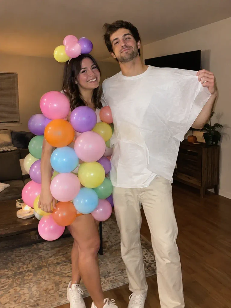 Candid iPhone-style photo of a couple in flowy fortune teller attire beside a round clear prop, standing at an indoor Halloween party, warm ambient lighting, genuine candid framing, realistic fabric texture, no text overlay