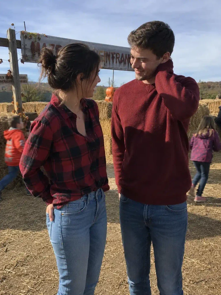 Candid iPhone photo of a couple both wearing denim overalls over simple long-sleeve tees, hers cream, his heather gray, denim fabric showing natural creasing at the straps and knees from crouching. They're crouched together reaching for a pumpkin on the ground, genuinely mid-motion, not posed, her laughing at something he said, his focus on the pumpkin. Real hand detail gripping the pumpkin stem. Background: rows of pumpkins on the vine, dirt ground, distant barn structure softly blurred. Bright natural daylight, real shadows across the dirt, slightly tilted candid framing with visible grain.