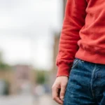 Description: A premium, realistic candid 21:9 website hero banner photograph featuring a stylish, modern men’s outfit idea, using a gray sweatshirt and jeans as the central focus. The image is captured with a natural, candid feel, with the subject positioned on the right half, leaving plenty of negative space on the left side for website text and CTA buttons. In the right-middle ground, a young man is photographed in a relaxed, candid pose while leaning casually against a textured concrete wall in an urban outdoor setting. He wears an oversized, heavyweight heather-gray crewneck sweatshirt with dropped shoulders and ribbed cuffs, paired with relaxed-fit, mid-wash blue denim jeans. The jeans have natural wear and a slight break over a pair of clean white sneakers. One hand is casually in his jeans pocket, while the other is by his side. His gaze is directed slightly away from the camera, giving a natural, lived-in feel. The outdoor natural daylight is soft and diffuse, highlighting the fabric’s rich textures—the heavy cotton of the sweatshirt and the rugged grain of the denim. The background features a soft-focus urban environment with blurred architecture, metal railings, and a hint of green foliage in the distance, providing a contemporary, city-vibe. The camera is at a medium-close cinematic perspective, bringing out the detail in the clothing while blurring the background with a shallow depth of field. The overall color palette is grounded in natural, cool tones—grays, blues, and urban neutrals—creating a highly aspirational and professional fashion banner. Photo captured on 35mm film, film grain, natural contrast. No text, logos, or UI.