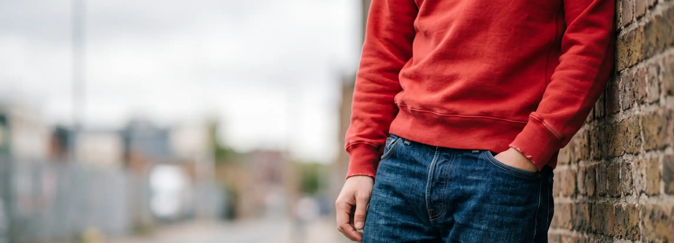 Description: A premium, realistic candid 21:9 website hero banner photograph featuring a stylish, modern men’s outfit idea, using a gray sweatshirt and jeans as the central focus. The image is captured with a natural, candid feel, with the subject positioned on the right half, leaving plenty of negative space on the left side for website text and CTA buttons. In the right-middle ground, a young man is photographed in a relaxed, candid pose while leaning casually against a textured concrete wall in an urban outdoor setting. He wears an oversized, heavyweight heather-gray crewneck sweatshirt with dropped shoulders and ribbed cuffs, paired with relaxed-fit, mid-wash blue denim jeans. The jeans have natural wear and a slight break over a pair of clean white sneakers. One hand is casually in his jeans pocket, while the other is by his side. His gaze is directed slightly away from the camera, giving a natural, lived-in feel. The outdoor natural daylight is soft and diffuse, highlighting the fabric’s rich textures—the heavy cotton of the sweatshirt and the rugged grain of the denim. The background features a soft-focus urban environment with blurred architecture, metal railings, and a hint of green foliage in the distance, providing a contemporary, city-vibe. The camera is at a medium-close cinematic perspective, bringing out the detail in the clothing while blurring the background with a shallow depth of field. The overall color palette is grounded in natural, cool tones—grays, blues, and urban neutrals—creating a highly aspirational and professional fashion banner. Photo captured on 35mm film, film grain, natural contrast. No text, logos, or UI.